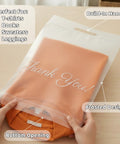 Hands holding a Frosted 10x13 Poly Mailer 100 Pack with Built In Handle Self Seal Matte Finish, containing an orange sweater.