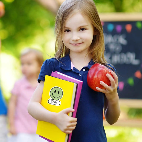 Child holding books with Smiley Name Tags Pack 120 Pcs Boho Pastel Smile Face Nameplates for Classroom, apple in hand.