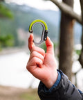 Hand holding HeroClip Small Carabiner Clip And Hook Three In One For Purses Strollers And Backpacks Stealth Black in outdoor setting