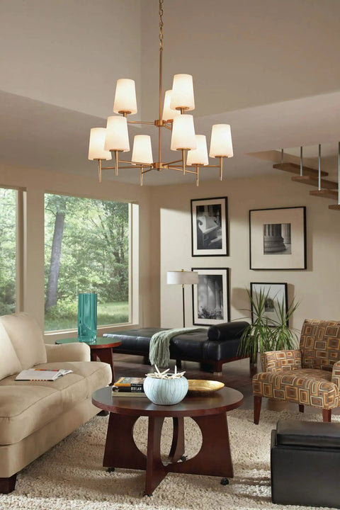 Living room with modern chandelier, beige sofas, framed art, and large windows overlooking trees.