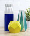 Geometric vases on a wooden table with a plant in the background, showcasing a blue, yellow, and teal color palette.