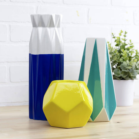 Geometric vases on a wooden table with a plant in the background, showcasing a blue, yellow, and teal color palette.