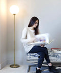 Luna LED Floor Lamp Dope Modern Tall Light For Living Rooms And Home Offices With Frosted Glass Globe Mid Century Style Standing Lamp In Brass Gold Finish Perfect For Reading In The Bedroom And Cozy Vibes next to a woman reading
