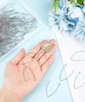 Hand holding stainless steel V shape earring hooks with a coin, part of 200 Pcs V Shape Stainless Steel Earring Hooks 19 Gauge Drop Ear Wires For Jewelry Making DIY Earring Findings Hoops In Handy Box Tarnish Resistant Durable Kit, hooks displayed with flowers.