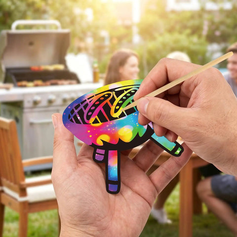 Hands using wooden stick on Camping Scratch Card Kit 24 Pack For Outdoor Parties 12 Unique Designs Revealing Rainbow Colors