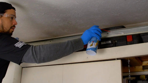Person applying Silicone Garage Door Lubricant Spray For Smoother Quieter Operation On Tracks Rollers And Hinges Will Not Attract Dust Or Dirt Tack Free Formula Reduces Friction And Noise So Your Door Roars Less And Moves Smoothly 9.3 Oz Case Of 12 on a garage track with gloves.