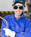 Man in welding safety glasses smiling, holding welding torch, wearing blue work suit with gloves, showcasing Kickass Welding Safety Glasses With HD Clear Lenses For Grinding And Outdoor Work Featuring Adjustable Nose Pads And Secure Ear Hooks For A Super Snug Fit That Gonna Shield Your Eyes From Sparks And Debris During Hard Job Sites In This Rugged 10 Pack Set.