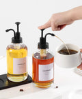 Hand using Two Pack Glass Syrup Pump Dispensers For Coffee Bar Setup With 84 Labels And Funnel, 16.9 Oz Each, Matte Black Pumps