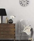 Modern living room with wood dresser, black lamp, decorative vases, transparent chair, and wall clock in simple interior design.