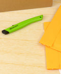 Westcott safety ceramic box cutter with green handle, side view on wooden desk beside orange envelopes