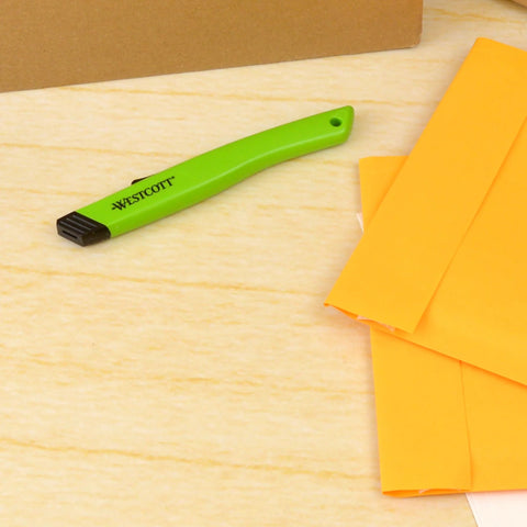 Westcott safety ceramic box cutter with green handle, side view on wooden desk beside orange envelopes