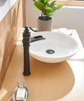 Oil rubbed bronze waterfall vessel sink faucet with single hole install, next to white basin on wooden vanity in rustic decor.