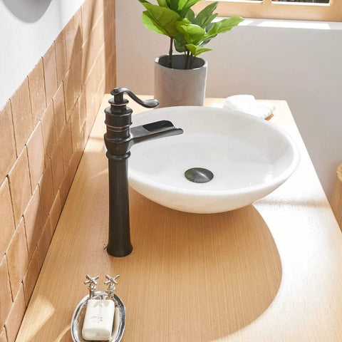 Oil rubbed bronze waterfall vessel sink faucet with single hole install, next to white basin on wooden vanity in rustic decor.