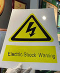 Person holding electric shock warning sign in yellow with symbol and text, displayed indoors.