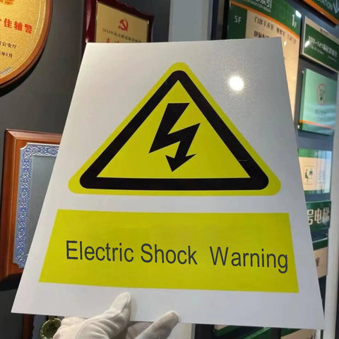 Person holding electric shock warning sign in yellow with symbol and text, displayed indoors.