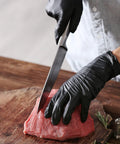 Black vinyl exam gloves; close-up of gloved hands slicing raw meat on a wooden cutting board.