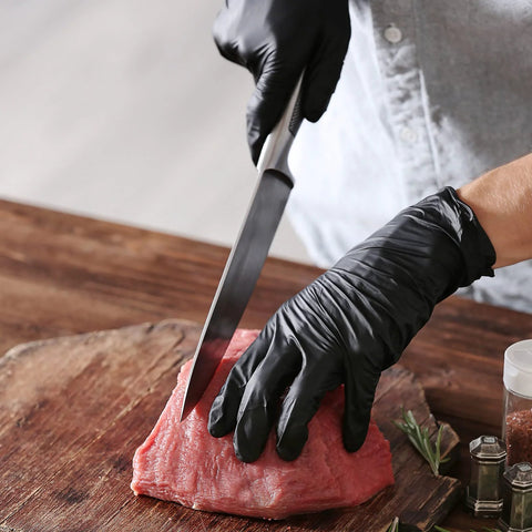 Black vinyl exam gloves; close-up of gloved hands slicing raw meat on a wooden cutting board.