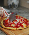 Person using cutter from Pizza Stone Set For Oven And Grill 15 Inch Round Cordierite Stone With 14 By 16 Inch Pizza Peel Stainless Steel Spatula And Cutter Complete 4 Tool Kit For Crispy Crust Bread And BBQ