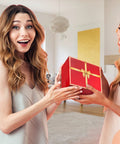 Two women exchanging a wrapped gift box with a gold ribbon in a bright living room.