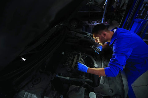Mechanic in blue uniform inspecting a car engine bay in a garage.