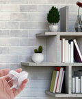 Hand holding transparent PVC corner guard for tables near shelves; 10 Pack PVC Corner Guards For Furniture And Steps Soft Corner Protectors For Safety Easy To Install Protect Edges Of Tables Walls And Cabinets Great For Baby Proofing And Kid Safe Homes visible in image