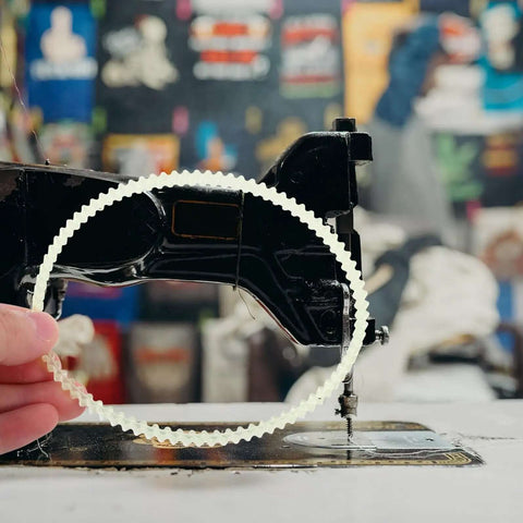 Person holding a rubber drive belt in front of a vintage sewing machine. Beef Up Your Sewing Setup With This 20 Pack Rubber Drive Belts For Sewing Machine Motors Replacement Parts For Household And Industrial Sewing Machines Pedal And Treadle Use Precise Fit Stable Transmission Quiet Operation.