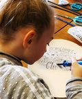 Round plywood craft board being drawn on by a child; light unfinished wood surface with scissors and markers on the table.