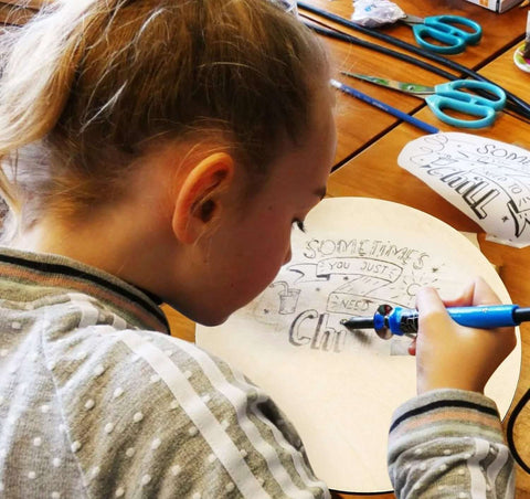 Round plywood craft board being drawn on by a child; light unfinished wood surface with scissors and markers on the table.