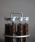 Eight Single Dose Espresso Bean Storage Tubes With Rotating Display Rack And Precision Funnel Set filled with coffee beans on a gray background.