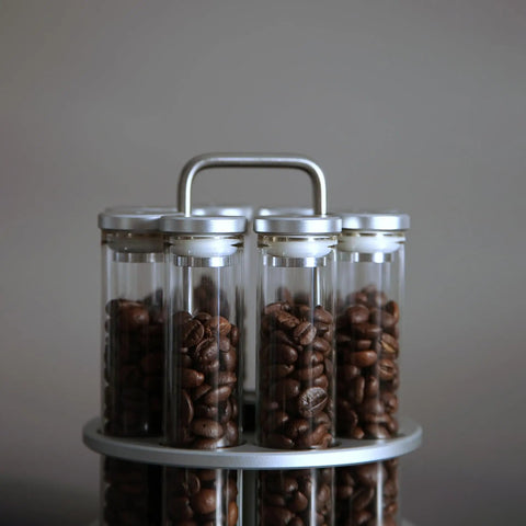 Eight Single Dose Espresso Bean Storage Tubes With Rotating Display Rack And Precision Funnel Set filled with coffee beans on a gray background.