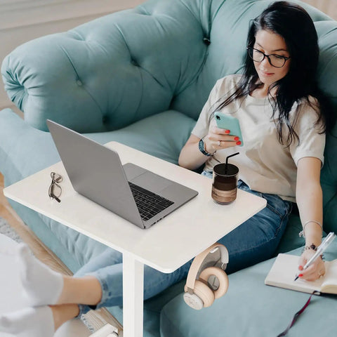 Mobile T Shaped Pneumatic Height Standing Desk on lockable wheels beside sofa with laptop, notebook, and headphones