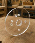Six Inch Clear Acrylic Router Base Plate With Precision Pre Drilled Holes For DWP611 And DCW600B Compact Routers on a wooden surface.