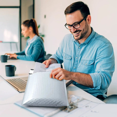 Man using Bubble Mailers 8.5x12 Inch White Pack Of 25, holding poly padded envelope at office desk, self-seal feature visible