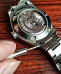 Close-up of stainless steel wristwatch with visible automatic movement; precision pin remover tool near the bracelet pin