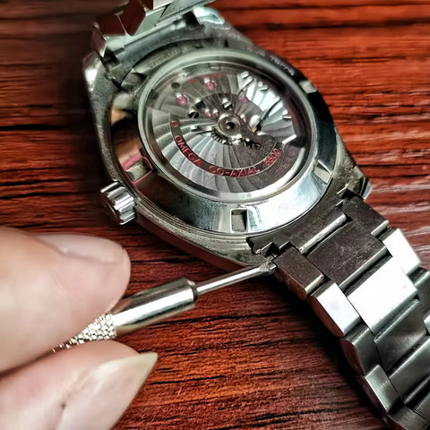 Close-up of stainless steel wristwatch with visible automatic movement; precision pin remover tool near the bracelet pin