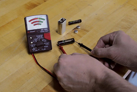 Testing batteries with Analog Battery Tester With Test Leads For All Common Batteries From 1.5V Button Cells To 12V Lantern Batteries, AA And AAA, 9V, 14 Battery Range, Easy Read Meter, Black on wooden surface.