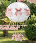 Hand holding pink bow hot air balloon centerpiece with 9.8 inch paper lantern, mini woven basket, and wooden sticks outdoors.