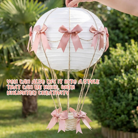 Hand holding pink bow hot air balloon centerpiece with 9.8 inch paper lantern, mini woven basket, and wooden sticks outdoors.