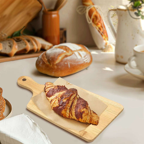 Bamboo cutting board with handle on a kitchen counter; croissant on parchment beside loaf.