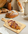Bamboo cutting board with handle, 11x5 in, on kitchen counter with parchment and croissant.