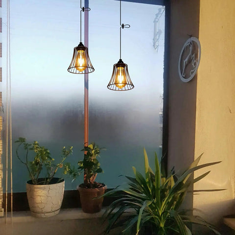 Two Three Pack Battery Operated Outdoor Chandelier Pendant Lights With Rustic Black Metal Bell Shade hanging near indoor plants.