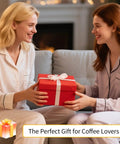 Two women exchanging a gift box, labeled as "The Perfect Gift for Coffee Lovers," on a cozy couch.