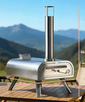 16 Inch Multi-Fuel Outdoor Pizza Oven For Wood Fired Or Gas Cooking Reaches 1112 Fahrenheit, Stainless Steel Portable Pizza Cooker For Parties, 2-In-1 System With Burners Sold Separately on a table with mountains in background