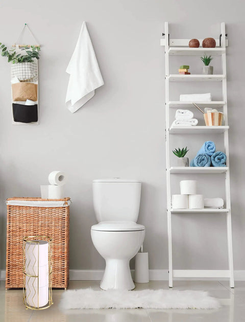 Bathroom setup featuring a modern toilet next to a wicker basket, ladder shelf with decor, and gold finish standing toilet paper holder.
