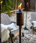 Two Pack 65 Inch Metal Black And Orange Outdoor Torches With Easy Install Lever for Patio with chairs in background