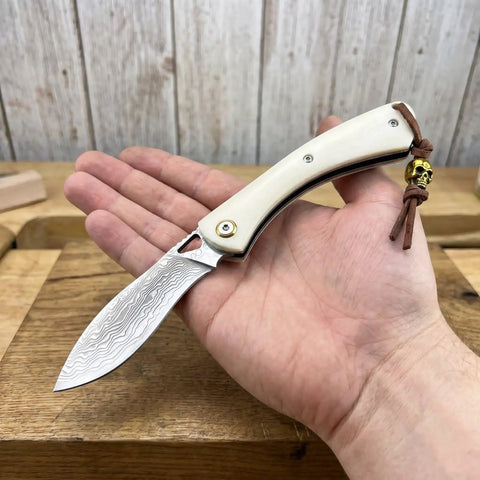 Folding Pocket Knife With 2.99 Inch Damascus Steel Blade And Ivory Synthetic Handle Ultra Portable EDC Knife For Camping And Daily Home Use Built For Everyday Carry And Outdoor Adventures Great Gift For Men, held open in hand, showing blade texture.