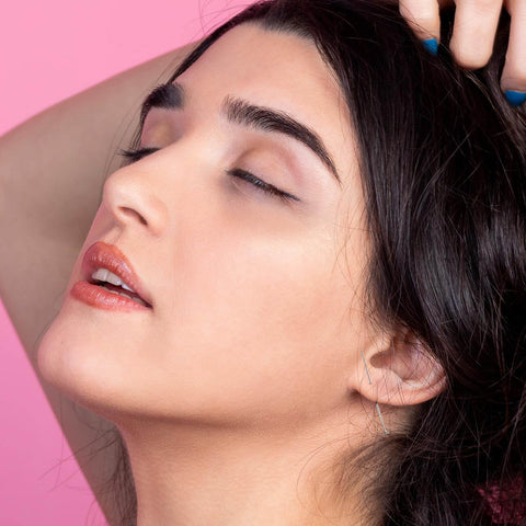 Side view of a person with dark hair, eyes closed, and subtle ear jewelry against a pink background.