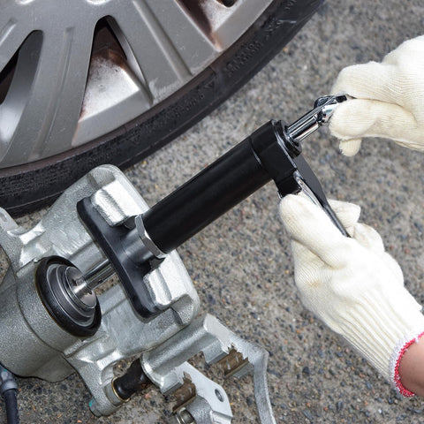 Side view of a pneumatic brake caliper piston tool in use on a car disc brake; black barrel, silver rod, gloved hand.