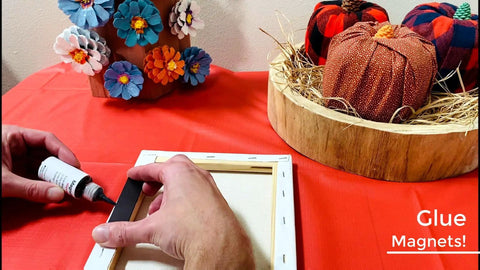 Person applying glue to attach magnets to a canvas on a table, near fall-themed decorations and orange fabric backdrop.