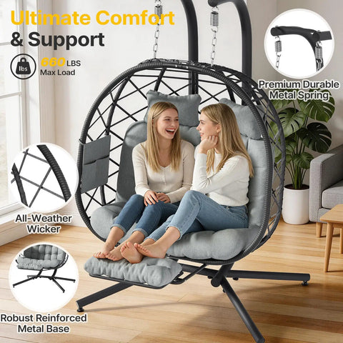Two people sitting in a Cozy Double Hanging Egg Chair For Two With Adjustable Footrest And Stand, Indoor Outdoor Wicker Swing With Cushions, 660Lb Capacity, Grey, in a living room.